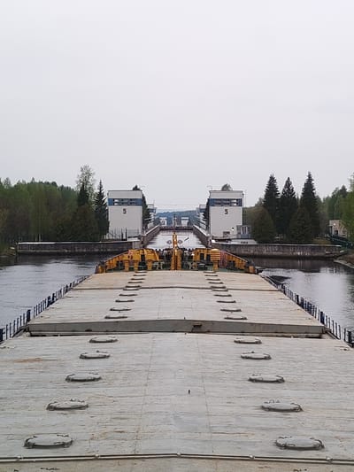 The image shows the Volga-Don Canal near Alekseevskoe, Russia. The view is from a large barge or ship, focusing on a lock system identified by the number "5" on the lock control towers. The foreground features the grey, flat deck of the vessel, with numerous circular access hatches. The canal is flanked by concrete walls and railings, with water on both sides. Background vegetation includes trees, and a small building is visible to the right. The sky is overcast.