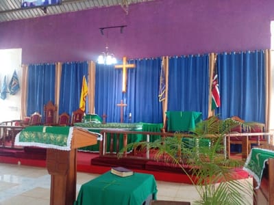 An interior scene shows a church altar area with a purple wall and blue curtains. A large gold cross is mounted on the wall behind the altar, flanked by flags. The altar itself is adorned with green cloths and crosses. Chairs are visible behind the altar, indicating seating for clergy. A wooden lectern stands in the foreground, covered with a green cloth. A potted plant adds a touch of nature to the solemn space.