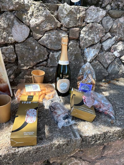 A collection of picnic items is arranged on a rough, stony ground, likely steps or an uneven outdoor surface, under bright, natural sunlight. No people are visible in the image.
The central subject is a dark green bottle of sparkling wine, identified by its label as "ABADIA DE MONTSERRAT CAVA". Its foil-wrapped cork is visible at the top. Surrounding the bottle are several packaged food items.
To the upper right of the Cava bottle is a gold-colored box labeled "1928" and containing "LLAMINETS CON TRUFA" (breadsticks with truffle), also noted as "LLAMINETS WITH TRUFFLE". Next to this box, in a clear plastic bag, are individual pieces of what appear to be dried sausages or cured meats. Another clear plastic bag contains cracker-like items, with a purple label bearing "Montserrat" and an illustration of a building, likely a monastery.
Below the Cava bottle, another gold box is visible, also marked "1928" and "SNACKS" by the brand "Jalgot". This box is for "SALCHICHÓN CON TRUFA" (salami with truffle). Nearby, a brown paper cup stands upright, and another similar cup is partially visible at the bottom edge of the frame. A small, dark, round item, possibly another piece of cured sausage, is in a plastic vacuum-sealed bag near the lower gold box.
The scene suggests an outdoor snack or picnic in a sunny location, consistent with the given context of Collbató, Spain. The strong sunlight casts distinct shadows, indicating a clear, bright day.