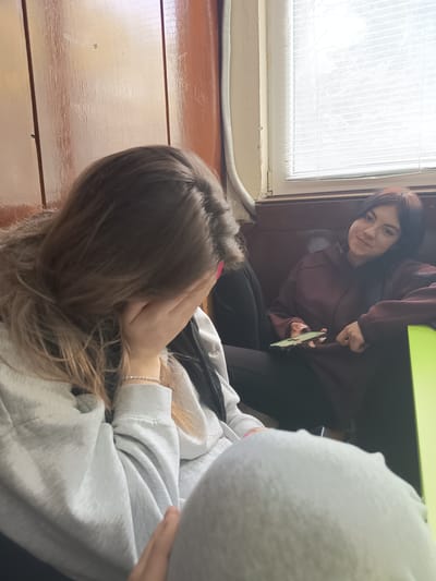 Two young women are sitting indoors. One woman is covering her face with her hand, while the other is looking towards the camera with a slight smile, holding a mobile phone. The environment appears to be an interior space with wood-paneled walls and a window with blinds in the background. The first girl is wearing a grey sweatshirt, a black puffer jacket and a white bracelet. The second girl is wearing a maroon hoodie and black pants. The photo appears to have been taken in Kavarna, Bulgaria.