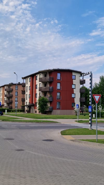 The image captures a daytime scene on a street in Sigulda, Latvia, likely in a residential area. Two apartment buildings are visible. The building in the foreground is a multi-story structure with a striking red and beige facade. It features multiple balconies with wooden railings. To the left, another apartment building, primarily beige and tan, is partially visible. The street in front is paved with grey interlocking bricks. A crosswalk is marked on the asphalt road that curves into the foreground. Traffic signs are present, including a yield sign, a pedestrian crossing sign, and a sign with a black and white striped pole. Green grass areas and trees surround the buildings and road. The sky above is blue with scattered white clouds.