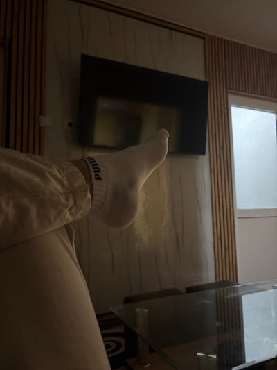 A low-angle, close-up shot depicts a person's lower leg and foot, wearing a white Puma sock, resting against a background featuring a wall-mounted flat-screen television and a window. The scene is interior, with artificial lighting creating shadows. The wall has a light marble-like pattern and horizontal wooden slats. A glass-topped table is partially visible on the right, with a patterned surface underneath. The image was taken in an indoor environment, presumably a residence. It is not possible to determine from the image alone if it was taken in Mirkovo, Bulgaria.