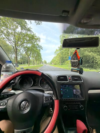 The image shows the interior of a car, specifically the driver's side, with a view looking out onto a road lined with trees. The steering wheel, dashboard, and center console are visible. The car is in motion, as indicated by the speedometer displaying a speed of 54 km/h. An air freshener and sunglasses hang from the rearview mirror. A bottle of "Erve Slenderbroek" water is placed in the driver's side storage area. The location is Lelystad, Netherlands, as indicated by the "U12 Lelystad" sign visible through the windshield. The time on the dashboard screen is 15:42. GPS coordinates 282365 and 52065 are visible on the car's digital display.