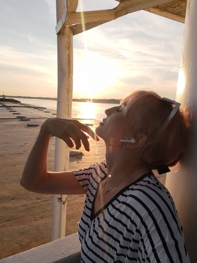 The image shows a woman with short, reddish-blonde hair, wearing a black and white striped shirt and white earbuds, leaning against a white cylindrical pillar. She has her head tilted back, looking upwards towards the setting sun. Her left arm is raised, and her hand is positioned near her face. Sunglasses are perched on top of her head. The scene appears to be outdoors, with a body of water and a distant shoreline visible in the background. The sky is a pale blue with wispy clouds, and the sun is casting a warm glow. The ground is a sandy or concrete surface with some visible lines. The overall impression is one of relaxation and enjoying the sunset. The location is specified as the city of Shchetinino, Russia.