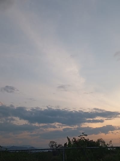 The image captures a serene evening sky over Parroquia Ocumare, Venezuela. The sky is predominantly a light, muted blue, fading into a warmer, orange hue towards the horizon. Several layers of clouds are present, with darker, more defined cumulus clouds closer to the horizon and lighter, more dispersed cirrus clouds higher in the sky. In the foreground, there's a line of green vegetation, including bushes and trees, partially obscuring what appears to be a chain-link fence. Distant mountains are faintly visible on the horizon.