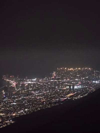 The image displays a wide, elevated view of a massive night gathering, with the crowd stretching horizontally across the frame. Thousands of people are present, their individual forms indistinguishable due to the distance and darkness. Their presence is primarily indicated by a multitude of small, bright lights they hold.

These lights appear as countless pinpricks of white, yellow, and occasionally red or blue, scattered densely across the elongated crowd. They are likely sources such as cell phone lights, flashlights, or candles. Faint outlines of structures, possibly buildings or other large infrastructure, can be discerned lining the path of the crowd on what would be the far side of the gathering. The background is predominantly dark, suggesting a night sky or unlit distant areas, with a subtle haze or cloudiness visible in parts.

The activity taking place is a large-scale collective event, possibly a demonstration, protest, vigil, or celebration, characterized by the participants' widespread use of personal lights. The sheer number of individuals and their coordinated illumination create a powerful visual impact. The time of day is clearly night. No discernible text is visible in the image. The location context provided is Alto Hospicio, Chile.
