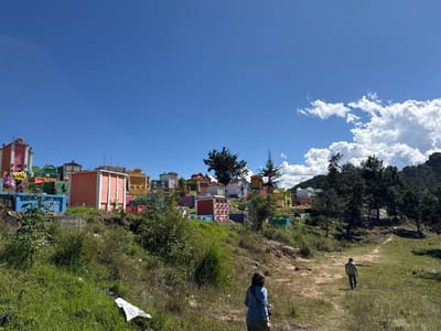 A colorful cemetery sits on a hill beneath a clear blue sky dotted with fluffy white clouds. The structures are painted in vibrant shades of orange, pink, yellow, and blue. Two people are walking along a path in the foreground, with grassy slopes and trees surrounding them. The scene evokes a sense of cultural vibrancy and peacefulness in a natural setting.