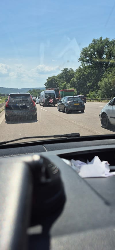 This media file shows a traffic jam on a sunny day, likely in Karlsruhe, Germany, given the location context. The scene is viewed from inside a vehicle, looking out at a line of cars, trucks, and a tractor. The time of day appears to be late morning or early afternoon, with bright sunlight casting shadows. The weather is clear and blue sky is visible with some scattered clouds.
The primary subjects are the vehicles caught in the standstill. These include a black hatchback with a Dutch license plate "37-KVS-5," a white van with a red and white striped safety sign, a green flatbed truck, and several other cars further down the line. A red tractor is also visible. The foreground of the image is dominated by the interior of the vehicle from which the photo is taken, showing a dark dashboard and what appears to be some papers or tissues on the passenger side.
The activity is clearly a traffic jam, with vehicles stopped or moving very slowly. There are no people prominently visible within the vehicles, though their presence is implied. The setting is a paved road, likely a highway or a major road, bordered by lush green trees and vegetation on one side, suggesting a rural or semi-rural environment. A hilly landscape is visible in the background.
Notable details include the license plates of some vehicles. One car in the foreground has a license plate "EL6 580CC." The overall mood conveyed is one of frustration or patience, typical of being stuck in traffic. The bright sunlight and clear sky might offer a slight contrast to the inconvenience of the traffic jam.
