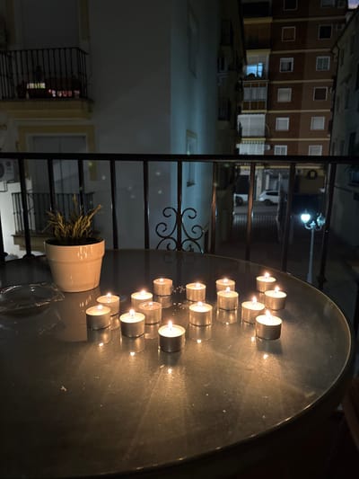 A group of lit tea light candles rest on a dark, possibly metal, round table. A white flower pot sits to the left, with wispy plants. A black iron railing divides the scene. Behind the railing, a multi-story building is visible with many windows. The scene is dark, illuminated primarily by the candle flames and an overhead street light. The image orientation is rotated 90 degrees counter-clockwise.
