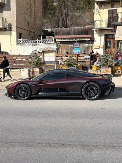 This image depicts an outdoor urban scene in Briançon, France, specifically adjacent to the Cafe Champ de Mars. The foreground features a dark metallic burgundy and black Aston Martin Valhalla sports car, parked facing left on a paved asphalt road. The vehicle exhibits multi-spoke black wheels and visible carbon fiber components on its lower body and side elements.

Behind the car, to the right of the frame, is the outdoor terrace of a cafe. A prominent sign above the establishment identifies it as "CAFE CHAMP DE MARS." The terrace is enclosed by a low wooden barrier and furnished with approximately seven yellow, wicker-style chairs and small square tables. Small potted evergreen trees are positioned among the seating arrangements. Seven individuals are observed seated at these tables, engaged in various social interactions. A menu board displays French text including "Gaufres" (Waffles), "Plats Chauds" (Hot Dishes), "Snack," and "Bière" (Beer). The cafe's building, partially visible, is a light yellow structure with dark brown window shutters and an upper-floor balcony.

To the left of the parked vehicle and forward of the cafe terrace, a woman walks barefoot towards the left side of the frame. She wears a dark top and light pants and is pushing a small, dark-wheeled stroller or buggy.

The background behind the cafe consists of a steep hillside densely covered with bare deciduous trees and some evergreens, suggesting a mountainous environment. The scene is illuminated by strong, clear daylight, casting distinct shadows.