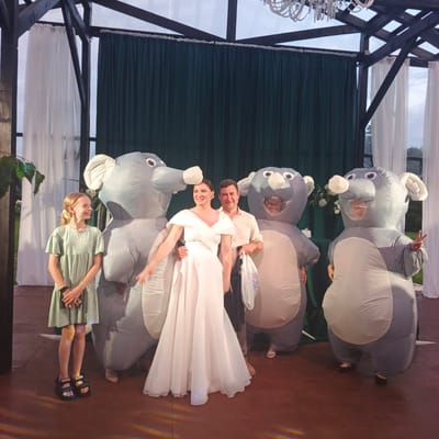 The image depicts a group of people posing in an outdoor event space, possibly a gazebo or open-air pavilion, in Svetloe, Russia. In the foreground, a young girl in a light green dress stands to the left, holding a small rectangular object. Beside her are two individuals wearing large inflatable elephant costumes. In the center, a woman in a flowing white dress and a man in a light-colored button-down shirt are positioned between the inflatable elephants. The man has his arm around the woman and holds a white plastic bag. To the right, another person in an inflatable elephant costume is visible. Behind the group, dark green curtains form a backdrop, with white sheer curtains draped on either side. The structure of the pavilion is visible at the top, with wooden beams and a translucent roof. A chandelier hangs from the ceiling. The ground is a reddish-brown color, likely a wooden deck or pavement. The overall atmosphere suggests a festive or celebratory occasion.