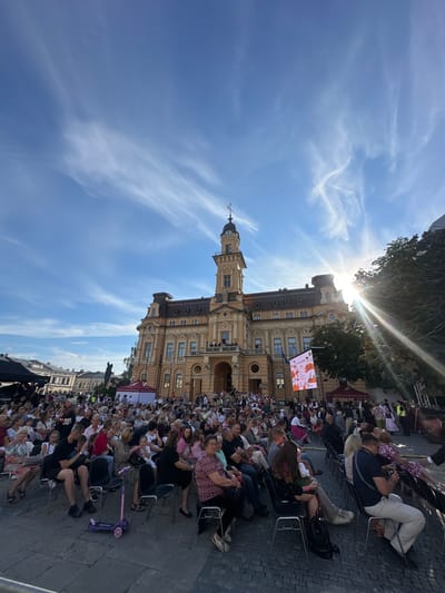 The image depicts an outdoor gathering in front of a large, ornate yellow building, identified as the City Hall in Nowy Sącz, Poland. A crowd of people, seated on chairs, are facing away from the camera, suggesting they are attending an event. The sun is shining brightly, casting lens flares and illuminating the sky, which features wispy clouds. Several tents are visible in the background, as is a large digital screen displaying an image. A child's scooter is parked on the cobblestone ground in the foreground. The architecture of the building, with its tower and decorative facade, indicates a historical and possibly important city center location. The atmosphere appears to be festive and social, with many individuals dressed in casual attire, some possibly in traditional clothing.