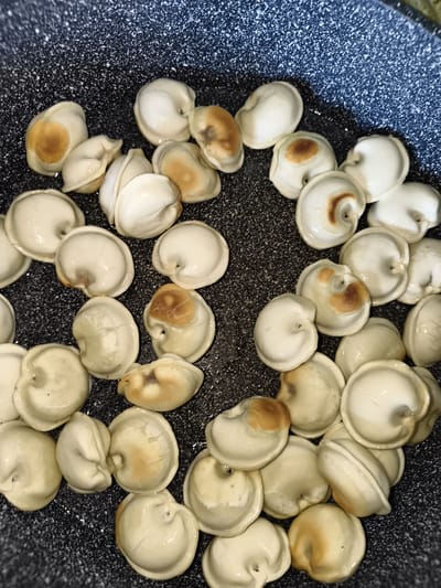 A close-up, overhead shot shows many pelmeni (dumplings) in a dark speckled pan. The pelmeni are light-colored, some with slightly browned areas from cooking. They are arranged in a scattered, somewhat overlapping pattern across the pan. The surface of the pan appears slightly greasy. There are no people or other objects visible in the image. The scene is a close-up of food being cooked.