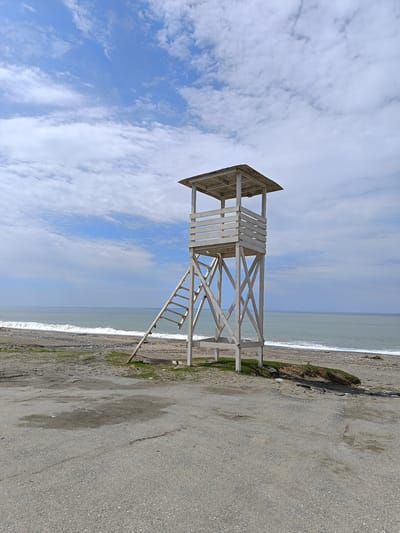The image shows a lifeguard tower on a beach. The tower is white, wooden, and elevated with a ladder for access. The beach is composed of sand and gravel. The ocean stretches to the horizon. The sky is blue with scattered white clouds. The photograph appears to be taken in Gulrypsh, Abkhazia. There are no people visible in the image. The environment is coastal.