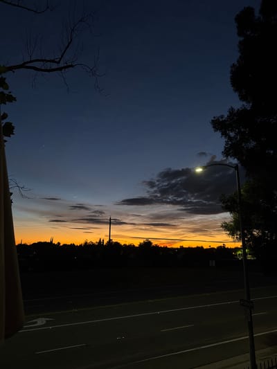 This is a high-angle, full shot of a street scene during twilight in Sacramento, California. The sky is a gradient of dark blue at the top to orange near the horizon, with scattered clouds. A streetlamp with its light on is visible on the right side of the frame, silhouetted against the sky along with a large tree. In the distance, the dark silhouettes of trees and buildings form a line across the horizon. A road with white lane markings is in the foreground, with a "LIBRARY" sign on a pole to the right. A bare-branched tree is silhouetted in the upper left corner, and a beige structure is on the far left edge of the image. No people are visible in the image.