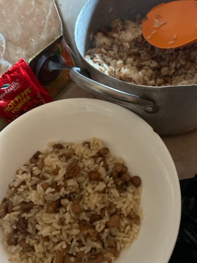 The scene, located in Egbe, Nigeria, depicts a prepared meal. In the foreground, a white bowl contains a serving of cooked white rice intermingled with brown beans. In the background, a metal pot holds a larger quantity of the identical rice and bean mixture, with an orange plastic spoon partially visible within the food. To the left of the pot, a red packet labeled "goodearth foods HOT PEPPE POWDER" rests on a light-colored, possibly tiled surface. A partially obscured yellow-brown packet is situated behind the red one. A dark, indistinct object is visible in the bottom right quadrant. No individuals are present in the image.