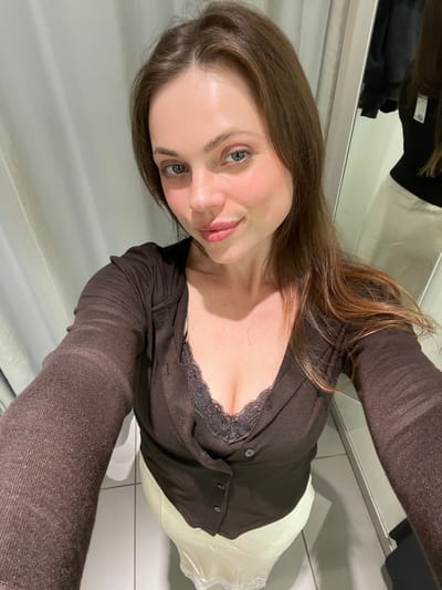A female individual is depicted taking a selfie in a modern indoor changing room. She has brown hair, light eyes, and is looking directly at the camera with a subtle smile. She wears a dark brown long-sleeved, buttoned top over a dark, lace-trimmed undershirt, paired with a light, possibly cream-colored, skirt or slip. Her arms are extended, holding the camera in a typical selfie pose. The background environment consists of a light-colored curtain on the left, a mirror on the right reflecting a dark hanging garment and portions of the room, and a tiled floor. This scene is characteristic of a contemporary retail or private indoor space, displaying no identifiable architectural or contextual elements corresponding to the city of Pompeii, Italy.