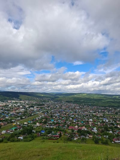 This is a high-angle, full-shot photograph of a densely populated town nestled within a valley. The town, identified as Sim, Russia, is spread across the frame, with a mix of multi-story buildings and numerous smaller residential houses with red, blue, and brown roofs. The houses are situated on relatively flat terrain, interspersed with trees and vegetation. A river or body of water is visible snaking through the town.
The town is surrounded by rolling, lush green hills and mountains, densely covered with forests. The landscape slopes upwards from the town towards the distant hills.
The sky above is a dynamic mix of white, puffy clouds and patches of bright blue sky, suggesting either a partly cloudy or a transitioning weather condition. The foreground features a grassy, undulating slope, indicative of a natural elevation from which the photograph was taken. The overall impression is a scenic view of a town in a rural, mountainous setting under a cloudy sky.