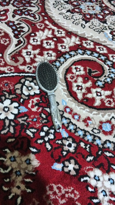 A hairbrush lies on a red and cream patterned carpet in an indoor setting. The carpet features a traditional floral design with accents of black, beige, and light blue. The hairbrush is oval-shaped with black bristles and a weathered, grey-toned handle that has a hole at the end for hanging. The lighting suggests it is daytime, and the image is taken from a slightly elevated perspective. There are no people visible in the image.