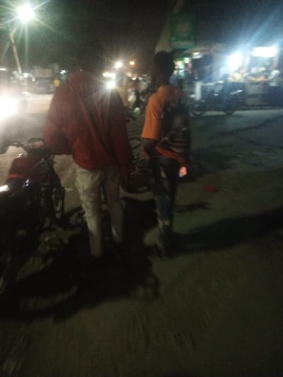 Two people stand at night, with one wearing a red jacket and light-colored pants, the other in an orange shirt and jeans. A motorcycle is visible nearby. The street is lit by bright streetlights, creating long shadows. In the background, there are other parked motorcycles and a brightly lit stall or shop. The scene is grainy and appears to be taken with a low-light camera.