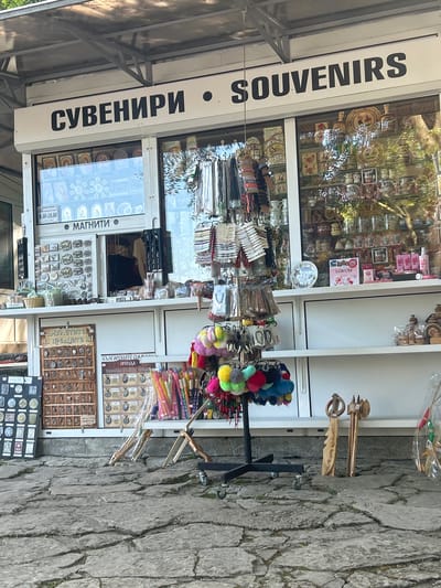 The image shows an outdoor souvenir stand in the city of Madara, Bulgaria. The stand features a white awning with the words "СУВЕНИРИ" and "SOUVENIRS" printed in black capital letters. Below the sign, a glass-fronted display showcases various items, including what appear to be icons, magnets, and other small trinkets.

On the left side of the stand, there are several display boards featuring magnets with different designs and historical motifs. A sign with text in Cyrillic script hangs on the left side of the display. In the center, a rotating rack is filled with colorful fluffy pom-poms and various beaded accessories. To the right of the rack, several wooden decorative items are displayed, including what resembles wooden sculptures and possibly keys.

The ground in front of the stand is paved with large, irregular stone slabs, with some grass growing in the cracks. The overall scene suggests a tourist-oriented marketplace or a small shop selling local crafts and souvenirs. There are no people visible in the image. The lighting indicates it is daytime.