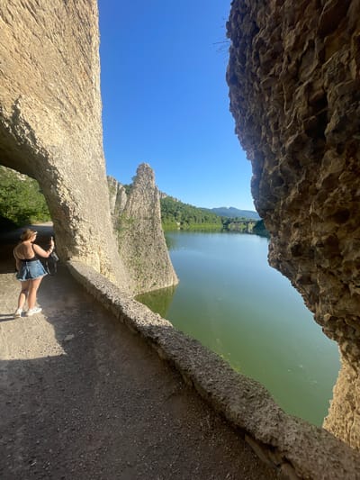In the city of Asparuhovo, Bulgaria, a woman stands on a stone pathway, taking a photograph with her phone. She is wearing a black tank top and denim shorts, with white sneakers. The pathway is carved into a natural rock formation that arches overhead on the left. Ahead, the pathway runs alongside a body of calm green water, bordered by a low stone wall. To the right of the pathway and water, more natural rock formations rise vertically. In the distance, across the water, a bridge spans the width of the frame, with hills and mountains visible beyond. The sky is clear and blue.