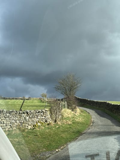 The image depicts a rural road scene under a heavily overcast, dark grey sky, characteristic of the countryside surrounding Lancaster, United Kingdom. The perspective is from within a vehicle, with visible reflections on a windshield, including faint partial text ("Good luck V" in the upper-right quadrant and "TIM" in the lower-right foreground).
A narrow, single-lane asphalt road, appearing worn with lighter patches of repair, curves sharply to the right and ascends gradually from the lower-center foreground into the mid-ground. The road is bordered on both sides by dry stone walls.
On the left side, a dry stone wall, partially covered with moss and lichen, defines the lower edge of a vibrant green, cultivated field that slopes upward. Further uphill in the mid-ground, a second dry stone wall runs parallel to the first, marking another field boundary. Several wooden fence posts are visible along these boundaries. Sparse, leafless bushes or small trees are situated along the field edges.
On the right side of the road, a continuous dry stone wall parallels the road's curvature as it ascends. Between this wall and the road, a strip of unkempt grassy verge contains dry, yellowed grass and small, bare shrubs. A singular, large, leafless deciduous tree with a dense branch structure stands prominently adjacent to the right-hand side of the curving road, silhouetted against the dark sky. A smaller, bare tree is visible further along the road on the same side.
The sky is dominated by thick, dark grey storm clouds, though a slightly lighter grey patch is visible in the upper-left quadrant. No human or animal subjects are present, and no specific actions or interactions are occurring. The lower-left foreground shows a partial view of a light-colored internal vehicle component.