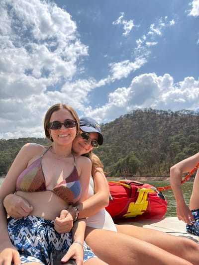 Two women pose for a photograph. One wears a bikini top with a multicolor triangle pattern and sunglasses. She has a necklace. The other wears a baseball cap and sunglasses. A life jacket is in the background. They appear to be on a boat. A body of water and forested hillside is in the background. This is likely on Lake Avándaro, Valle de Bravo, Mexico.