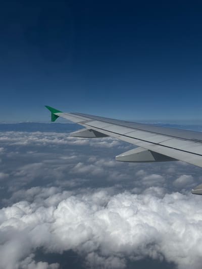The image shows an aerial view of an airplane wing with a green tip, extending over a landscape of clouds and mountains. The sky is a deep blue, indicating a clear day. The wing is white with dark gray panel lines, and a portion of the airplane's fuselage is visible. Below the wing, fluffy white clouds stretch across the horizon, obscuring the ground below. In the distance, a range of mountains can be seen peaking through the clouds. The scene appears to be during daylight hours, likely in the afternoon given the lighting. The perspective suggests the photo was taken from a passenger's window seat on a flight. There is no visible text in the image.