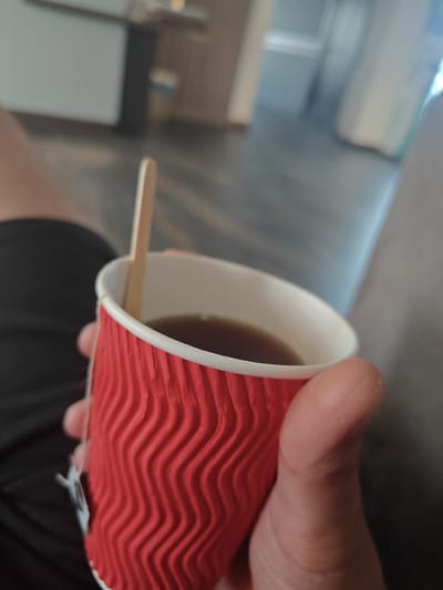 A person's hand holds a red paper coffee cup containing a dark liquid and a wooden stirrer. The cup has a distinctive corrugated texture. The background is blurred, showing muted tones of gray, brown, and white, suggesting an indoor environment. The person is wearing black shorts or pants.
