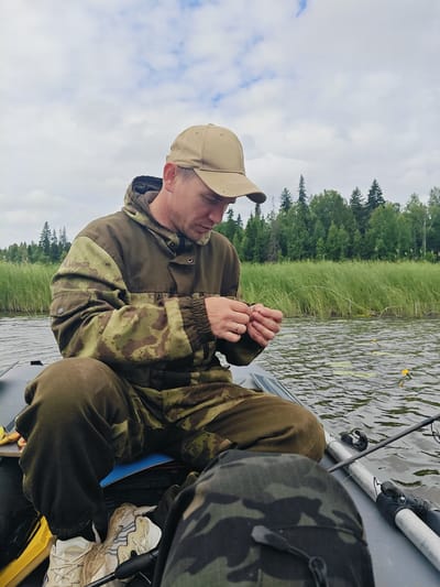 A man wearing a camouflage jacket and a tan baseball cap is sitting in an inflatable boat on a body of water. He is focused on his hands, which are holding fishing line and appear to be tying a knot. The boat is on a lake or river, with tall reeds and trees visible in the background. The sky is overcast with clouds. Fishing rods are present in the boat, and yellow fishing floats are visible in the water. The man is wearing sneakers. The image captures a moment of preparation for fishing.