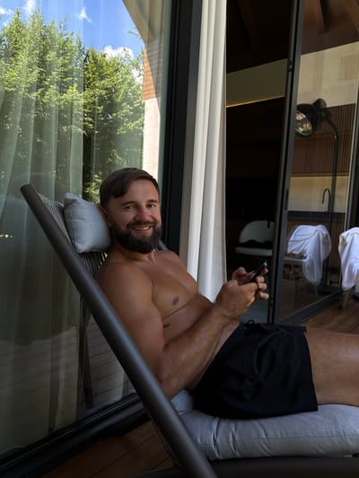 A man with a beard, shirtless, wearing black swim trunks, is reclining in a lounge chair on a wooden deck outside a building. He is holding a smartphone in his hands and looking at the camera with a smile. The background includes a large glass door or window reflecting trees and the sky, as well as interior elements such as a lamp, chairs, and draped towels. The scene suggests a resort or hotel setting, possibly in Bakovići, Montenegro.