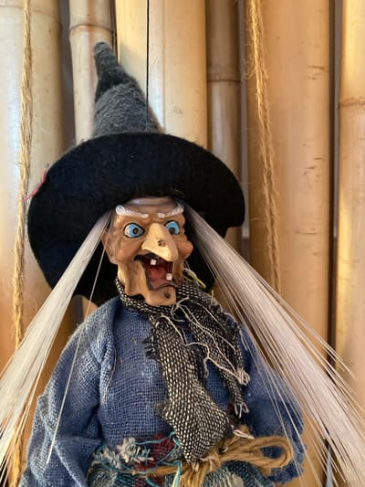 A doll, resembling a witch with a grotesque face, is displayed against a backdrop of bamboo poles. The doll wears a black pointed hat, a blue tunic, and a textured black and white scarf. White, thin fibers emanate from the doll, suggesting some sort of magical effect or a decorative element. The doll's face is detailed with blue eyes, an open mouth revealing teeth, and wrinkled skin. The overall impression is one of a handmade craft or decoration, possibly for a holiday like Halloween. The scene is likely indoors, given the controlled lighting and setting. No text is visible in the image.