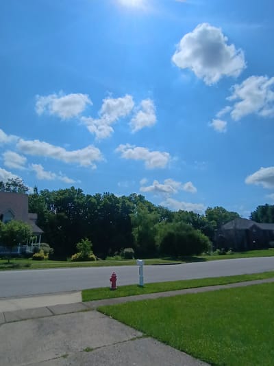 This is a sideways, eye-level, full shot of a suburban street on a clear day. The sun is shining brightly on the left side of the frame, casting rays across a vibrant blue sky dotted with fluffy white clouds. The street itself is a light gray asphalt, with a neatly trimmed green lawn on the right and a sidewalk made of gray concrete. A bright red fire hydrant stands out on the grassy verge. Further down the street, a light blue mailbox is visible. Beyond the lawn, a dense line of lush green trees forms a backdrop, with hints of houses peeking through the foliage. The overall impression is one of a peaceful and sunny day in a residential area. There is no visible text in the image.