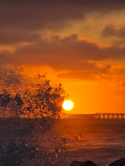 The image depicts a sunset scene at the Oceanside Pier in Oceanside, California, USA. The sun is a bright orb low on the horizon, casting a warm orange glow across the sky and illuminating the clouds. In the foreground, a wave crashes, creating a spray of water that catches the light. The Oceanside Pier extends into the ocean, with silhouetted figures visible on it, likely enjoying the sunset. The water is rippled, reflecting the colors of the sky. The overall atmosphere is serene and picturesque.