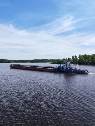 The image displays a blue and white tugboat actively pushing a long, unladen cargo barge, predominantly reddish-brown with blue accents, from the right side of the frame towards the left. The vessels are situated on a wide expanse of dark blue-grey water, exhibiting numerous small ripples and a distinct wake trailing behind the tugboat. The distant shoreline across the water is densely covered with a continuous stretch of lush green forest, composed of both deciduous and coniferous trees, indicating an undeveloped natural environment. The sky above is a bright, clear blue with scattered, delicate wispy white clouds. No individuals are visible on the vessels or along the distant banks. The visual characteristics of a large, undeveloped waterway surrounded by extensive forest are not consistent with the urban landscape of Mitino, a residential district within Moscow, Russia.
