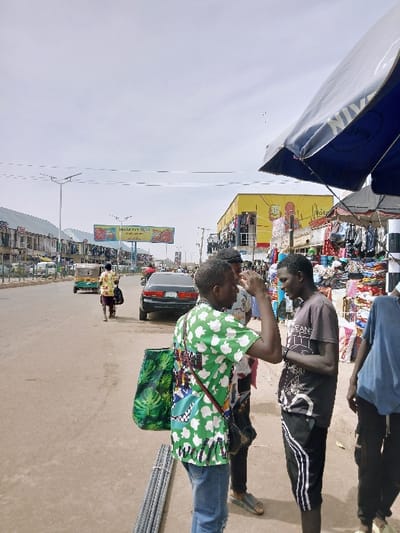 The image depicts a busy street scene in Kwekwe, Zimbabwe, under a bright, overcast sky. In the immediate foreground, a bundle of metal rebar rests on the ground. Three males stand on the right side of a paved road. The central figure wears a green shirt with white and multicolored patterns, blue jeans, and carries a dark green leaf-patterned tote bag over his left shoulder. His right hand is raised to his temple. He faces a second male to his right, who wears a dark t-shirt with white lettering, dark shorts with white side stripes, and dark sandals. A third male is partially visible on the far right, wearing a light blue t-shirt and dark trousers.
In the midground, a dark sedan is parked or moving slowly on the road, facing away from the viewer. Further down the road, a green and yellow auto-rickshaw (tuk-tuk) is visible, followed by a person walking away from the camera. The road surface is light brown and appears dry.
Along the left side of the street, a row of multi-story buildings with numerous windows extends into the distance. On the right, large commercial structures are present, including a prominent bright yellow building adorned with red graphic elements, likely a retail establishment. Immediately to the right of the foreground subjects, market stalls are set up under a large blue and white umbrella, displaying various clothing items. Overhead power lines or communication cables traverse the sky. Streetlights line the road in the distance.