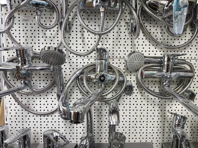 The image displays a retail display of chrome bathroom fixtures, specifically shower heads and faucets, suspended against a white pegboard with black dots. The display features multiple complete shower sets, each with a faucet, hose, and shower head, as well as individual faucet components. The overall impression is one of a product showcase in a store or showroom.
Several pieces of visible text provide product information, including labels with text such as "BATERIE DUS", "ZERIX FAD", and price information like "$50.00" and "$650.00". These labels suggest that the items are for sale.
The lighting appears to be typical of an indoor retail environment, with bright, even illumination that highlights the reflective surfaces of the chrome fixtures. The arrangement of the items is orderly, with each shower set or component hung or placed for optimal viewing. The setting is clearly a commercial space, likely a hardware store, bathroom supply store, or a section within a larger retail establishment in Cricova, Moldova. There are no people visible in the image. The time of day and weather are not discernible from the photograph. The overall mood is neutral and informative, focusing on the presentation of the products.