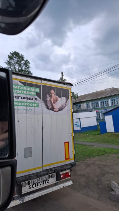 The image depicts the rear and side of a white truck with a trailer in what appears to be an outdoor, possibly semi-rural setting. A billboard on the trailer shows a woman lying down and text in Russian, which translates to "We invite drivers" and a phone number. Below the billboard, the truck's rear bumper displays a license plate reading "BE 8934 16 RUS" and the brand name "SCHMITZ". In the background, there are buildings, including one that appears to be a wooden house with blue trim and another smaller blue and white building. Power lines are visible against a cloudy sky. The location is identified as Nizhny Ingash, Russia.