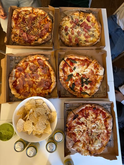 The image shows a high-angle shot of a table laden with food and drinks. There are five pizzas in cardboard boxes. The pizza toppings include ham, mushrooms, cheese, tomatoes, and basil. Next to the pizzas is a large white bowl filled with tortilla chips and a container of guacamole. Five cans of beer are scattered near the chips. Portions of people wearing jeans are visible in the upper left and lower left corners. The boxes contain newspaper print. The scene is in an indoor setting with a white table. It is not possible to determine if it is in Burjassot, Spain.
