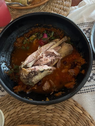 The image displays a partially consumed meal served in a dining setting within Guanajuato City, Mexico. The central subject is a black, round ceramic bowl containing a dish primarily consisting of a large portion of shredded cooked meat, possibly lamb or poultry, with visible bone fragments. This meat is submerged in a reddish-brown, semi-liquid sauce that contains dark, bean-like particles and lighter, soft vegetable matter. The dish is garnished with small green herb sprigs and several vibrant pink flower petals scattered on the sauce. The inner rim of the bowl exhibits dried residue from the sauce. The bowl rests on a light brown woven placemat. In the upper left background, a clear glass holds a pink beverage. To the right, a white fabric with a dark patterned border is visible, and further back, a portion of a light brown ceramic plate with a wrapped food item and a silver fork can be seen.
