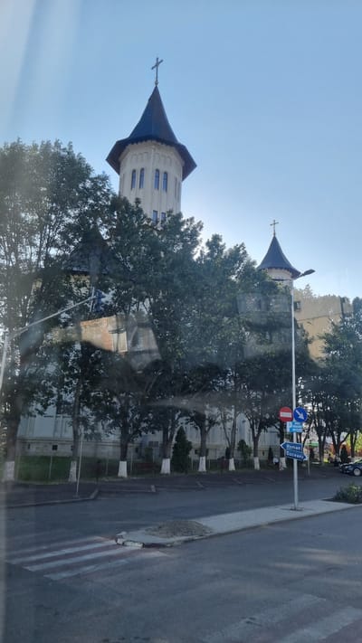 The image shows a view of the city of Fălticeni, Romania, focusing on a large building with two prominent towers, likely a church or monastery. The building has a white facade and dark, pointed roofs topped with crosses. Trees with white-painted trunks partially obscure the building. In the foreground, there is a street with a pedestrian crossing. Traffic signs are visible, including one pointing left with "Roman" and "Suceava" written on it, indicating directions to these cities. A blue sign above it reads "ZONA CU PLATA" which translates to "paid zone". A dark-colored car is parked on the right side of the street. The sky is clear and blue, suggesting it is daytime. There are no people clearly visible or engaged in any significant action. The scene is static and captures a moment of urban landscape.