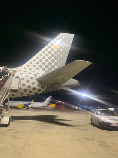 At night on an airport tarmac in Sant Boi de Llobregat, Spain, a Vueling Airlines airplane is parked. Its tail, decorated with a pattern of gray polka dots and a yellow circle, is prominently featured in the foreground. A set of mobile stairs is attached to the side of the aircraft, with several people ascending and descending. In the background, another Vueling airplane and a yellow and red structure are visible. A white van, with the license plate WB 32167, is parked on the tarmac to the right of the frame, illuminated by the aircraft's lights. The scene is lit by various airport lights, creating reflections and lens flares.