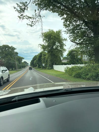 The image shows a view from inside a vehicle driving down a two-lane road in Upper Southampton Township, USA. The road is lined with trees and green grass. On the right side of the road, there is a white vinyl fence. A speed limit sign indicating 40 MPH is visible. In the distance, there are other vehicles on the road and houses partially visible through the trees. The sky is partly cloudy. The interior of the vehicle is also visible in the foreground, including a dashboard and windshield with a heads-up display showing speed information.