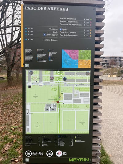 The image displays a large, dark grey outdoor information board, serving as a map and directory for the "PARC DES ARBÈRES" in Meyrin, Switzerland. The board is positioned outdoors, with a light-colored wooden fence visible behind its top edge. The background shows bare-branched trees, suggesting a winter or early spring season, and indistinct modern buildings under an overcast sky.

The information board is divided into several sections. On the left, a column lists destinations like "Rue des Arpenteurs," "Rue des Coopératives," and "Esplanade des Récréations," each with approximate walking times (e.g., "1 min," "2 min," "3 min") and directional arrows. Further down, categories such as "Vestiaires," "Stade," "P Sports," "P Centre Sportif," "Place de la Diversité," and "Parc de la Découverte" are listed, also with time estimates. A smaller, colorful inset map in the upper left corner shows a broader neighborhood overview, divided into colored zones.

The main right section of the board features a detailed, light green map of the park, highlighting various facilities including sports fields (labeled A, B, C, D, E, F, G), buildings like "ÉCOLE DES VERGERS," and paths. A prominent red "VOUS ÊTES ICI" (You are here) marker indicates the viewer's current location on the map. The park's name, "PARC DES ARBÈRES," is also visible on the map itself.

On the far right, a vertical section displays various legends and icons under headings such as "ACTIVITÉS NATURE," "PISTE CYCLABLE," "POINTS D'INTÉRÊT," "ÉQUIPEMENTS ET SERVICES," and "ACTIVITÉS SPORTIVES ET LUDIQUES." These legends use symbols to represent features like toilets, play areas, and picnic spots. The emergency number "112" is prominently displayed next to a phone icon, along with symbols for accessibility, cycling, and a bus stop (indicated by "10"). The name "MEYRIN" is clearly visible in green text on the far right. No people are visible, and the scene depicts a quiet, functional informational setting during daytime with overcast weather.