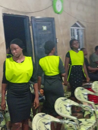 Three young women stand wearing black dresses and bright yellow vests. They are in a room with white walls and a green clock. There is a dark doorway and a small window covered with metal bars. The room is lit by overhead lighting. In the foreground are stacked white chairs with blue-grey patterns. There are several people sitting behind them in the background.