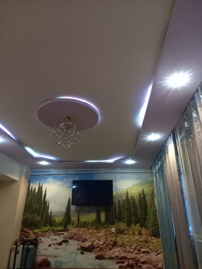 The image shows a living room with a scenic wallpaper of a forest and river. The room features a modern ceiling with recessed lighting and a chandelier. A television is mounted on the wall. The overall impression is that of a contemporary, well-lit living space. The location is described as being in Saruu, Kyrgyzstan.