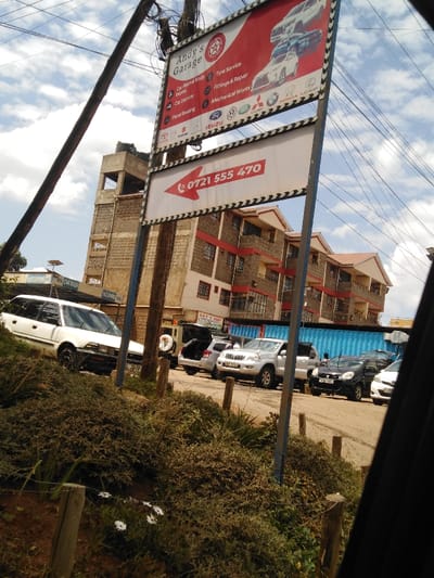 The image displays a commercial street scene in Kikuyu Township ward, Kenya, under a partially cloudy sky. A large, prominent vertical sign for "Andy's Garage" dominates the center, featuring a red and white design listing services such as Car Paint & Body Work, Tyre Service, and Mechanical Works, along with logos of various car manufacturers (e.g., Toyota, VW, Honda, Mercedes). Directly below this, a smaller white sign with a red left-pointing arrow displays the contact number "0721 555 470."

To the left, a multi-story building constructed from light-colored concrete blocks, topped with a water tank, is partially visible. To the right and behind the main sign, another multi-story residential or mixed-use building with multiple balconies, red trim, and a reddish-brown tiled roof is situated. Utility poles with numerous overhead wires are visible on the left and framing the central sign.

The foreground consists of dense, unkempt green and brown vegetation with a few white daisy-like flowers. Vehicles are parked on an unpaved or dirt surface. A white Toyota Corolla station wagon (partial license plate K... 359P) is visible on the far left. In the mid-ground, a silver Toyota Land Cruiser Prado SUV (partial license plate K... 178X) is parked, its tailgate open. Further back, several other vehicles, including a black car and a white car, are partially obscured. A large, light blue shipping container is visible in the background, likely serving as a workshop or storage unit. One individual is faintly visible near the blue container, and another appears to be near the open tailgate of a silver SUV. The scene indicates an active garage and parking area.