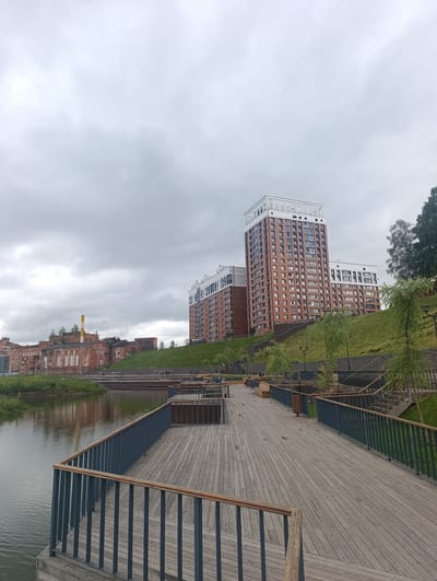 The image depicts a view of the embankment area of the city of Naro-Fominsk, Russia, along the Desna River. In the foreground, a wooden boardwalk with a dark metal railing stretches out, leading towards a body of water on the left. The boardwalk is empty, suggesting a quiet, possibly overcast day.
Across the water, on the left side of the image, are older brick industrial buildings, characterized by their red brick facades and large windows. One of these buildings has a tall, yellow-capped chimney. These structures appear to be a former industrial complex, now likely repurposed or undergoing renovation.
On the right side of the image, the landscape transitions to a modern residential area. Two tall, multi-story brick buildings with white and glass elements rise prominently, set against a grassy slope. These buildings are a stark contrast to the older industrial structures, indicating a mix of old and new development in the area.
The sky is overcast with heavy, gray clouds, contributing to the subdued lighting of the scene. Several trees with wispy branches are scattered along the embankment, some closer to the boardwalk and others further up the slope near the modern buildings. Streetlights are visible along the pathway, and a few benches are placed on the boardwalk and seating areas on the slope, hinting at public use. There are no people clearly visible in the immediate foreground or middle ground. The water reflects the muted colors of the sky and the surrounding architecture.