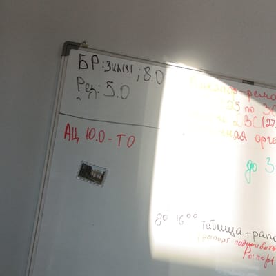 The image displays a white dry-erase board mounted in an indoor setting in Hlybokaye, Belarus. The board is positioned at an angle, tilting upward from left to right. A strong, diagonal glare from a bright light source significantly illuminates and obscures the right half of the whiteboard.
On the upper left portion of the board, two lines of text are written in black marker: "БР: ЗАКЛ; 8.0" and "Ред: 5.0". Below this, in red marker, is the text "АЦ 10.0-ТО". Directly beneath the red text, slightly left of center, a small rectangular object resembling a postage stamp is affixed. This object depicts a historical building, possibly a church or a palace, characterized by a prominent tower or spire.
The right side of the whiteboard is largely washed out by the glare, making much of the text illegible. However, fragmented words and numbers in various colors—yellow, orange, green, and red—are discernible. These include segments such as "...ремон...", "25 по 30", "...BC(27)", "...ная орг...", and "до 3..." in the upper and middle right sections. Towards the bottom right, below the glare, additional text written in black and red markers is visible: "до 16°°", followed by "Таблица + рано...", and a parenthetical phrase "(рапорт подписать)", with "Ронпорт" below it. No individuals are present in the scene.